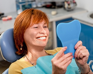 A middle-aged woman looking at her smile in the mirror after receiving dentures