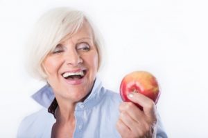 Woman with white hair wearing a light blue shirt is looking at an apple and smiling